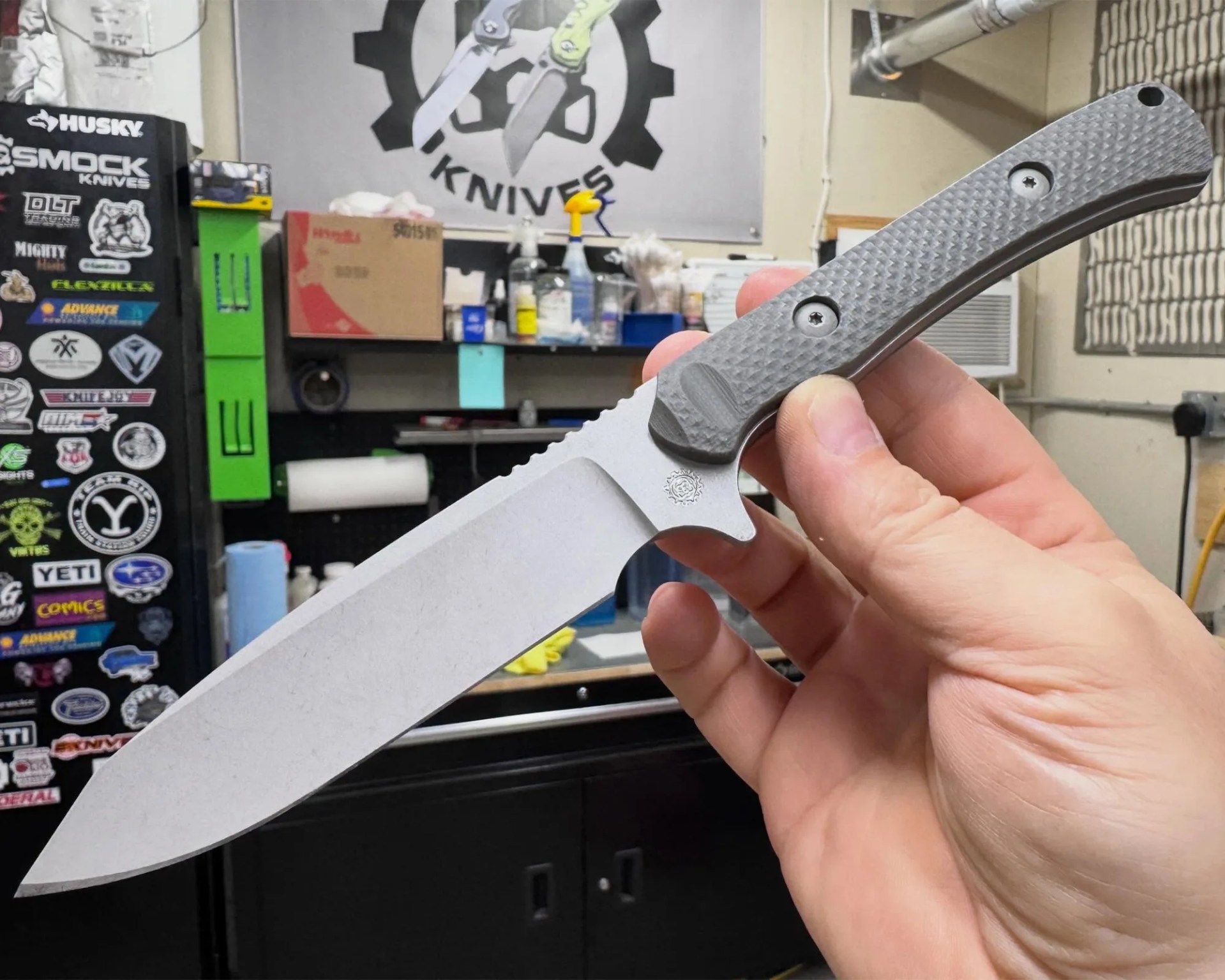 Hand holding a large fixed-blade knife with a textured gray handle in a workshop setting.