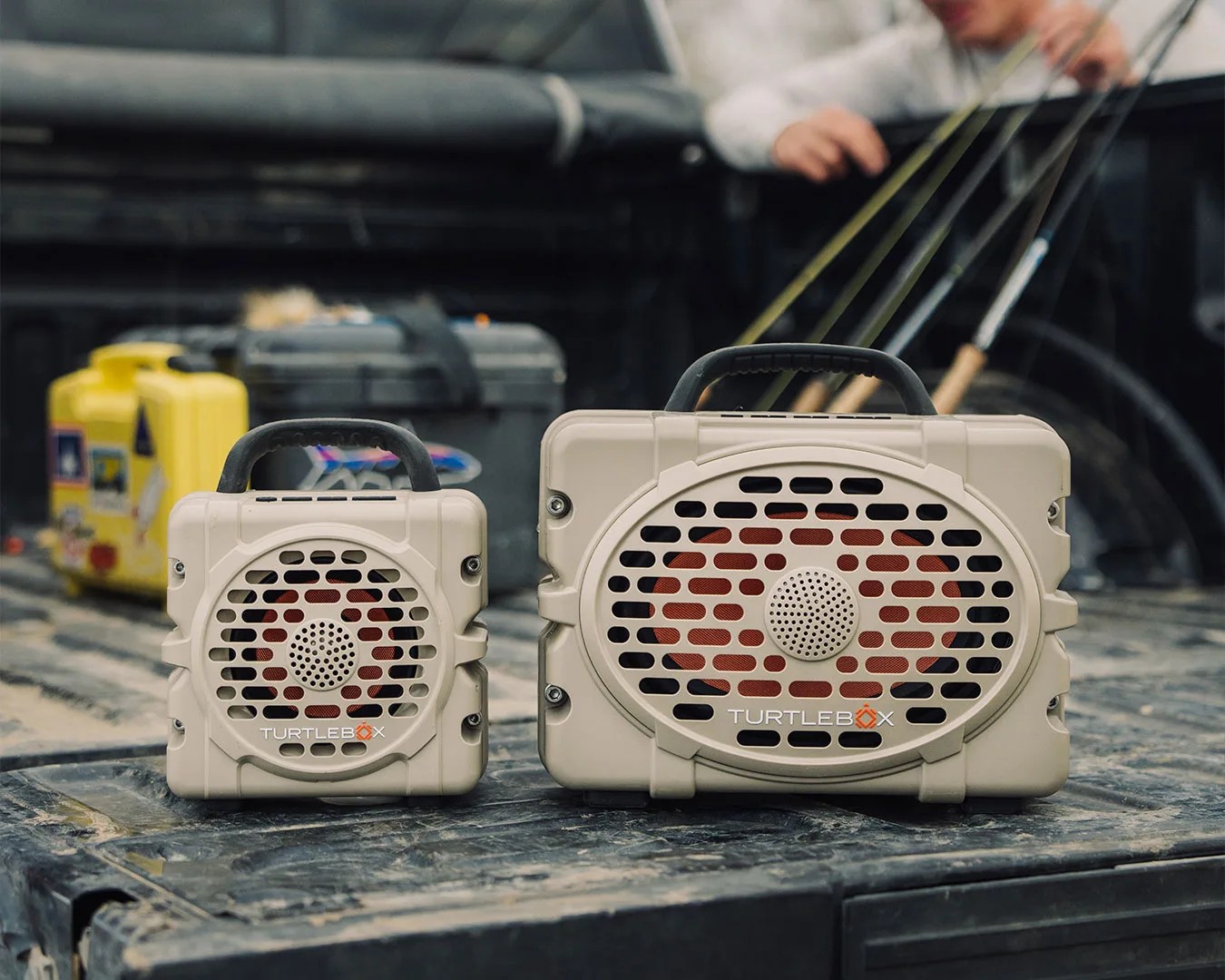 Two beige Turtlebox portable speakers with black handles on a dusty truck bed with fishing rods in the background.