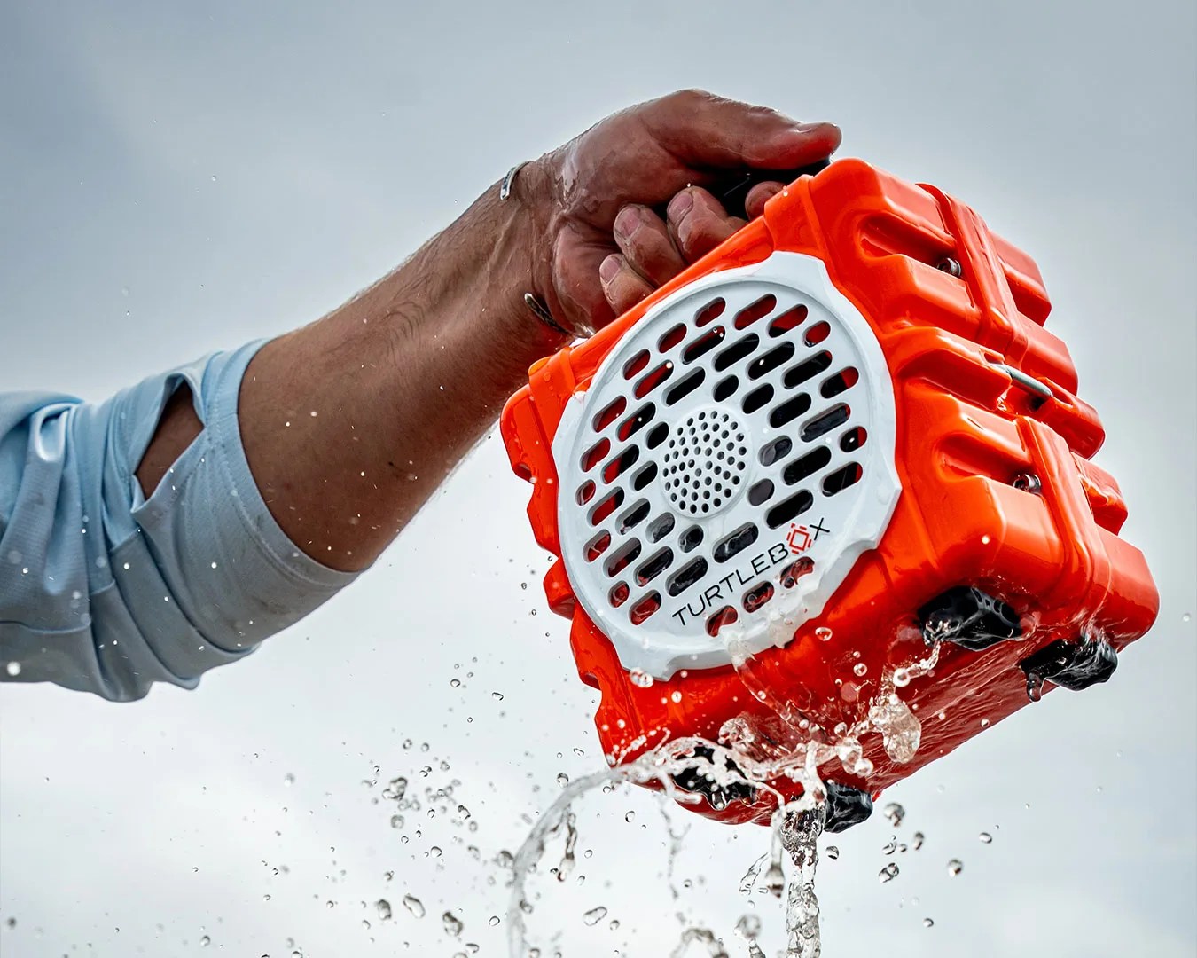 Hand holding an orange Turtlebox waterproof speaker with water splashing around it.