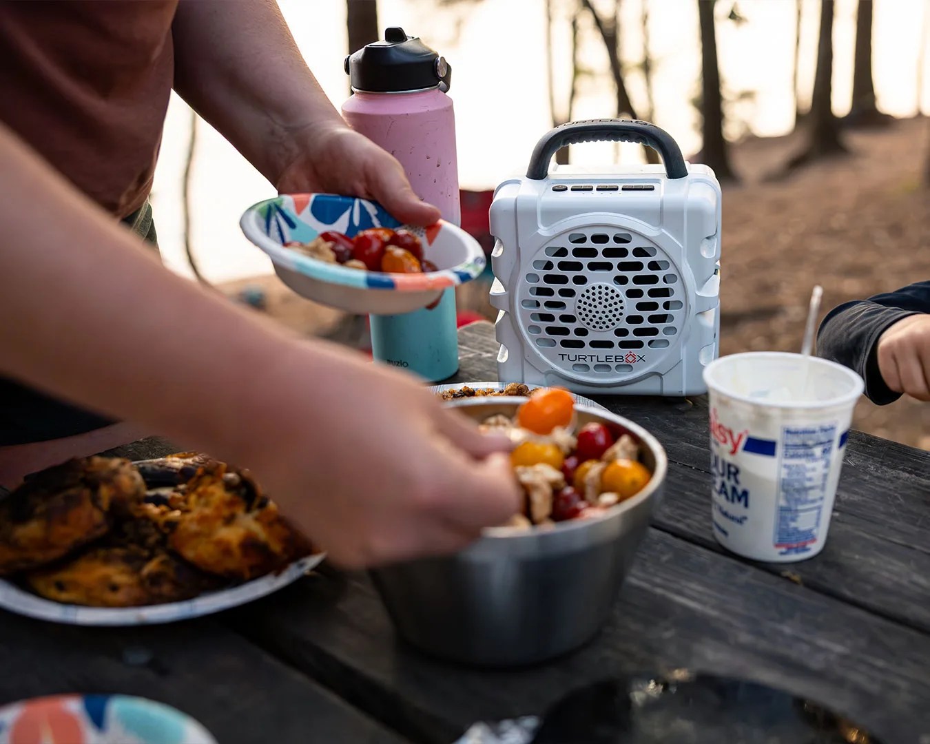 White Turtlebox portable speaker with black handle on a picnic table next to a pink water bottle and food bowls.