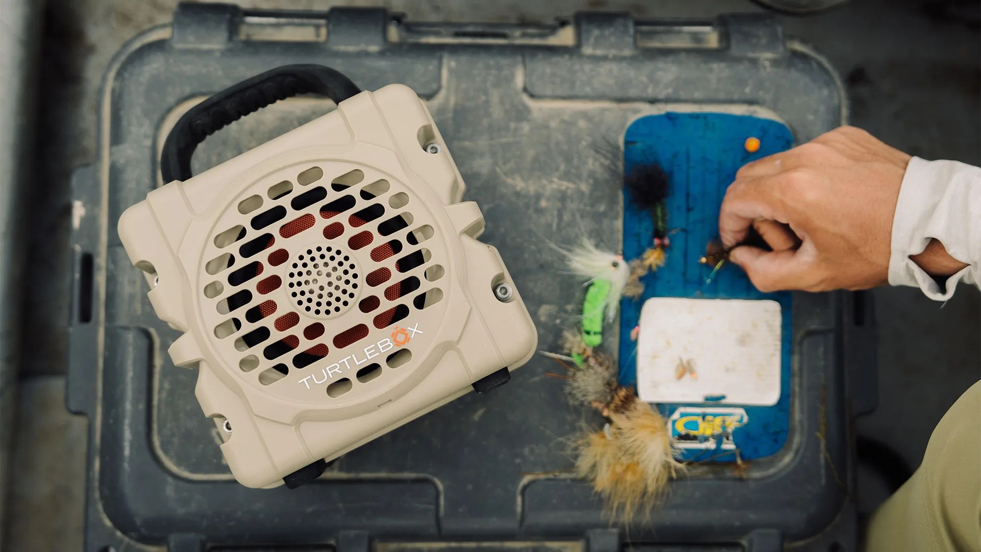 Beige TurtleBox speaker with black handle on a dark plastic case next to a hand selecting fishing flies on a blue board.
