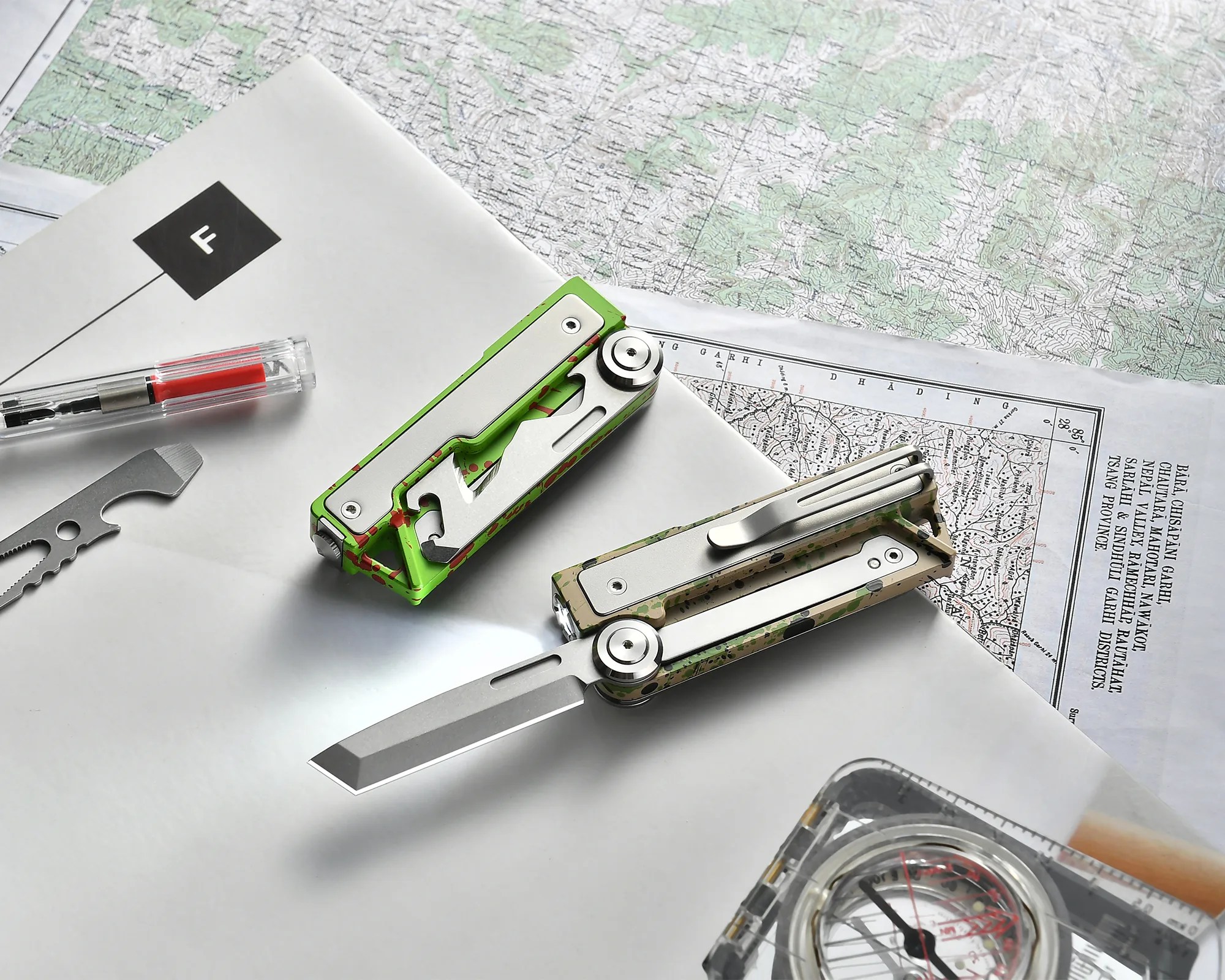 Two camo-patterned folding knives with silver handles on top of maps, alongside a multi-tool and a compass.