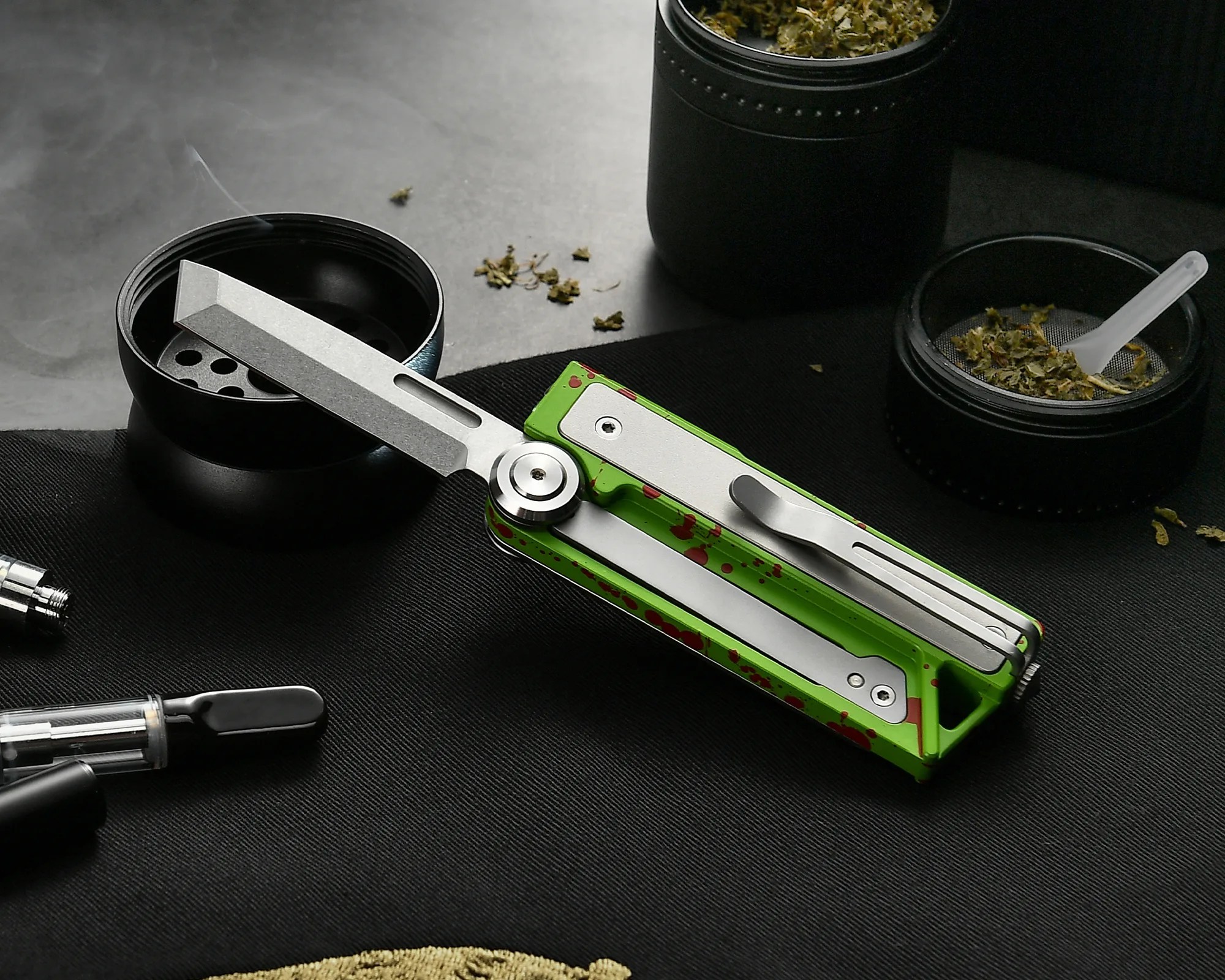 Green and silver butterfly knife with red splatter pattern resting on a black surface near black containers with dried herbs.