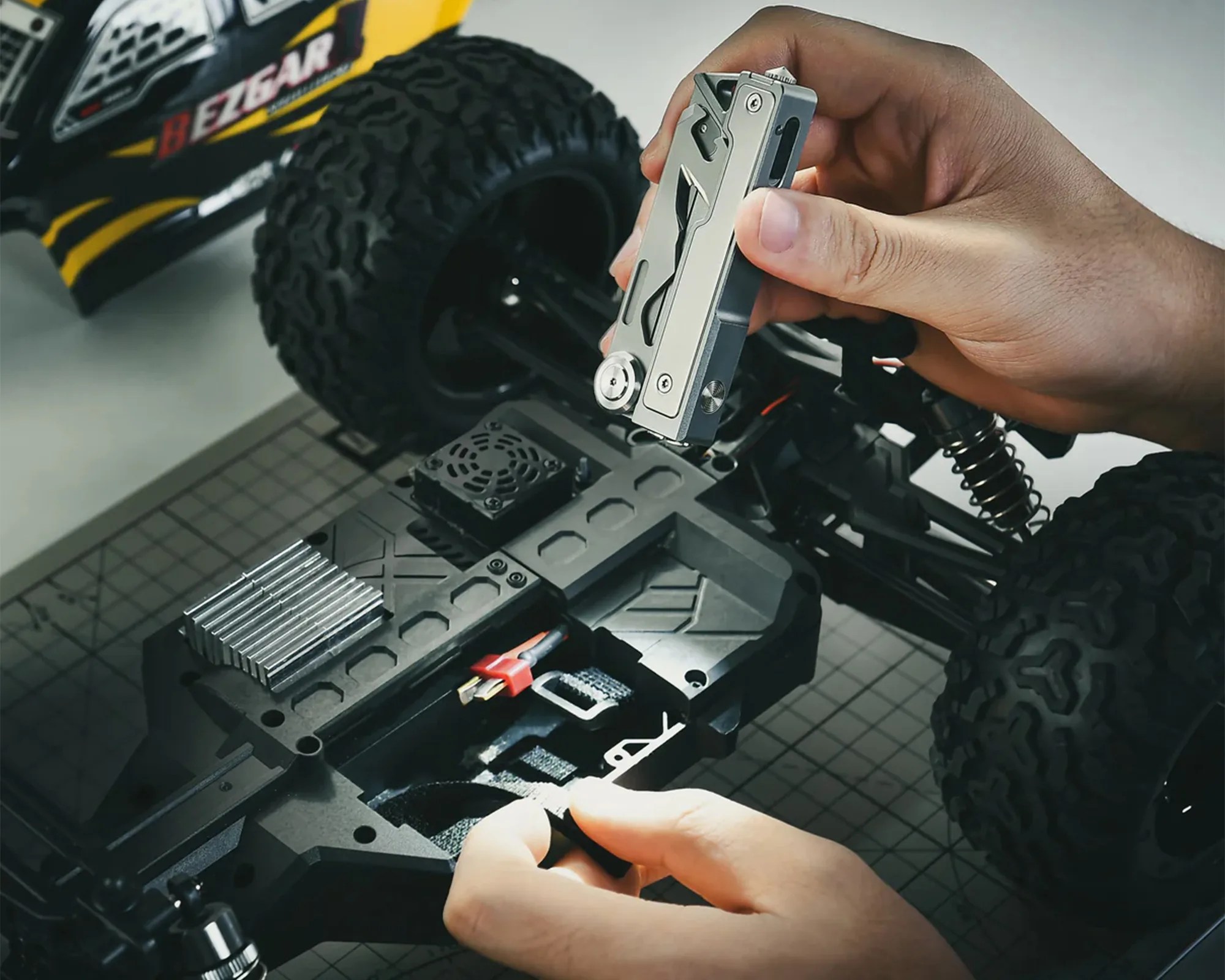 Hand holding a silver multi-tool above the chassis of a black and yellow RC car.