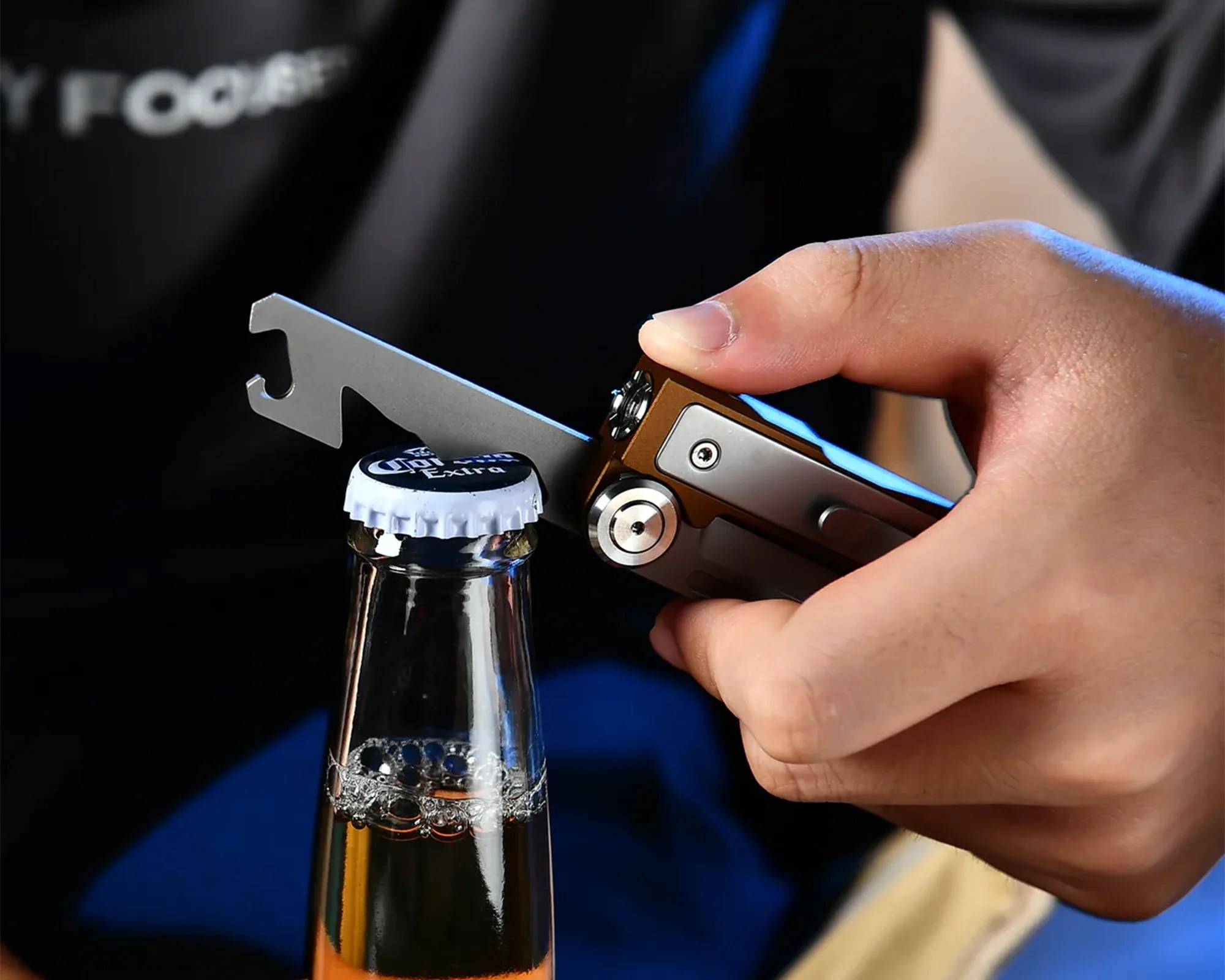 Hand holding a multi-tool bottle opener removing the cap from a Corona Extra beer bottle.