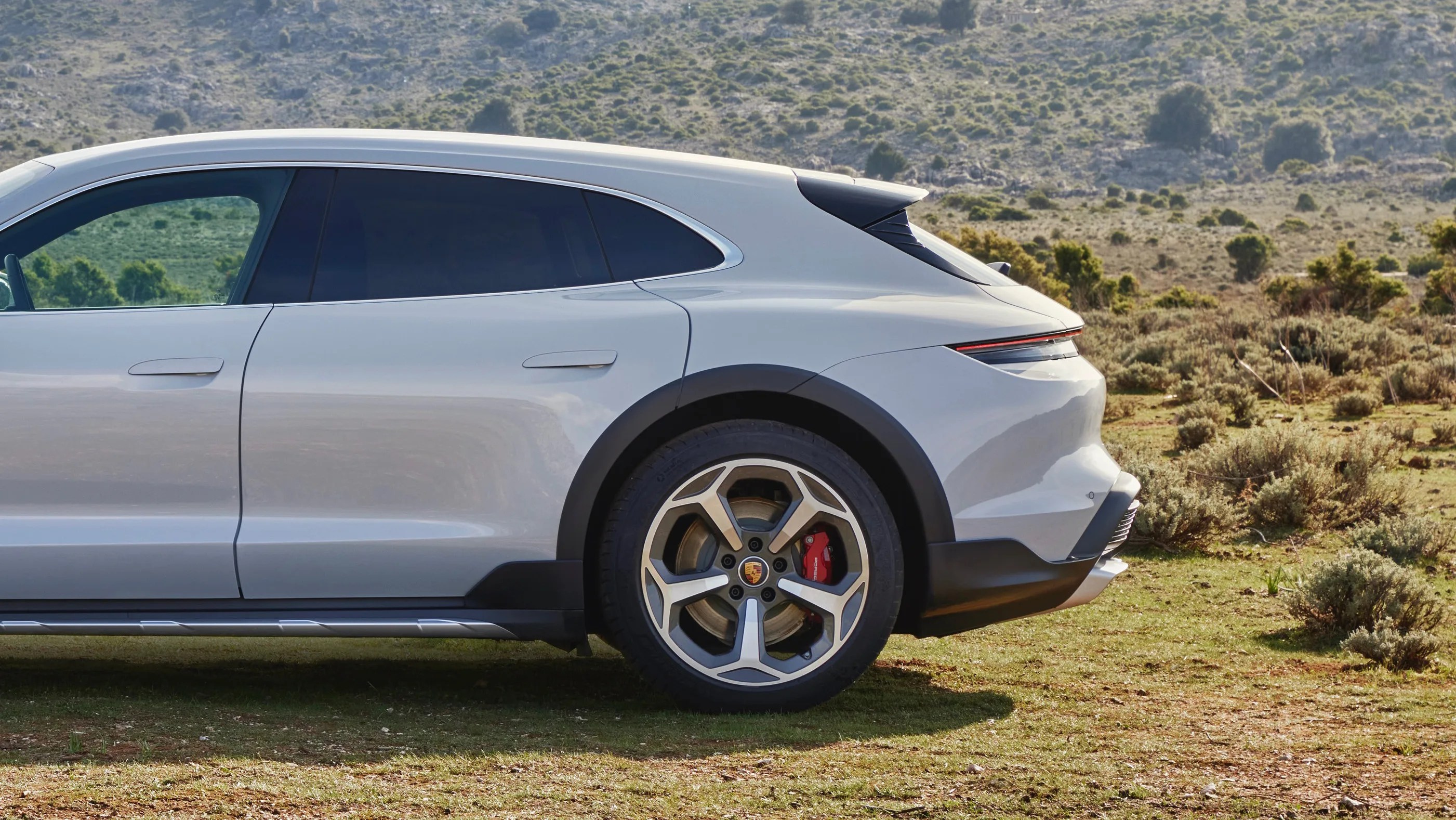 Silver Porsche electric SUV rear side with black wheel arches and red brake calipers on a grassy terrain.