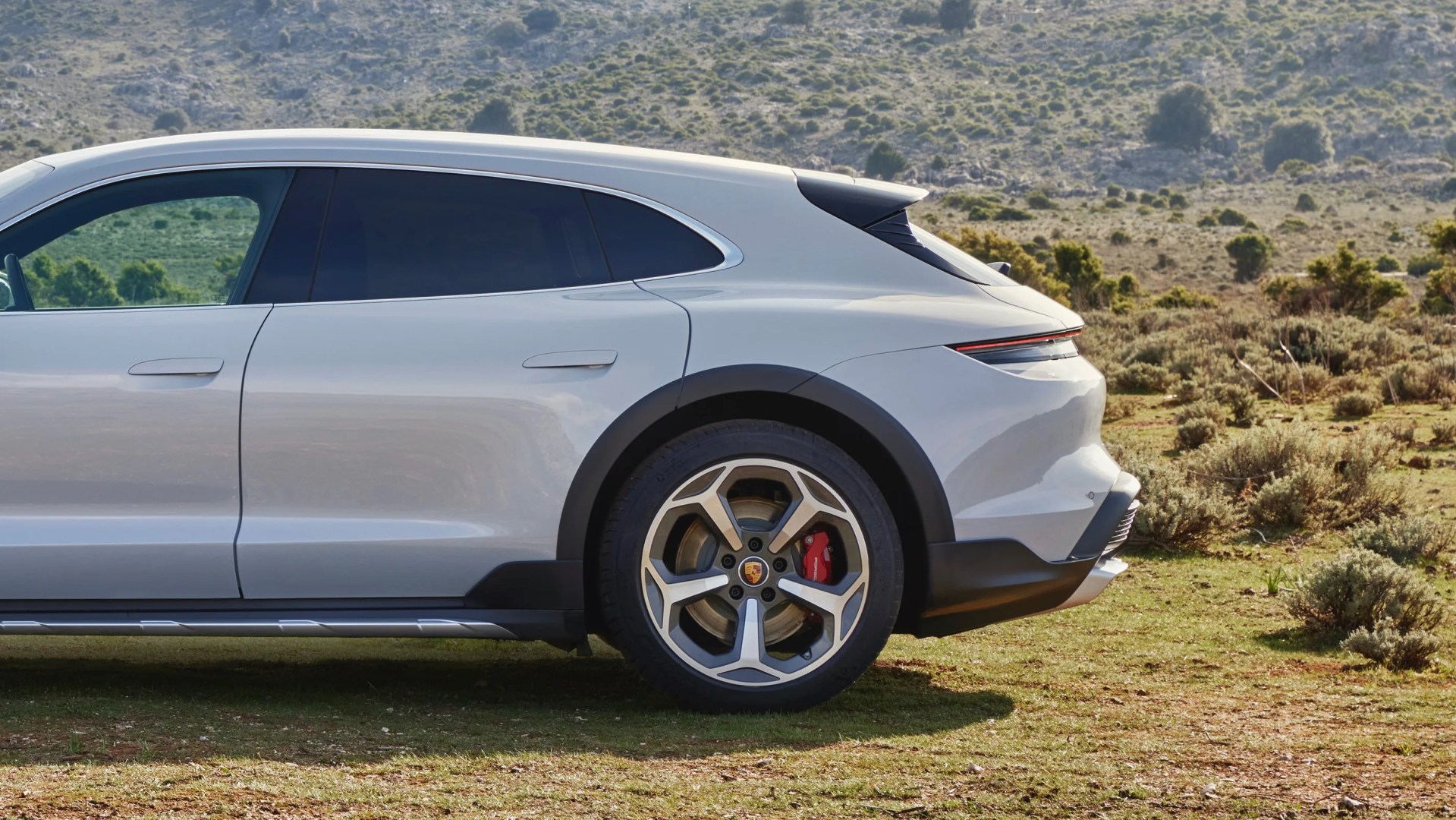 Silver Porsche electric SUV rear side with black wheel arches and red brake calipers on a grassy terrain.