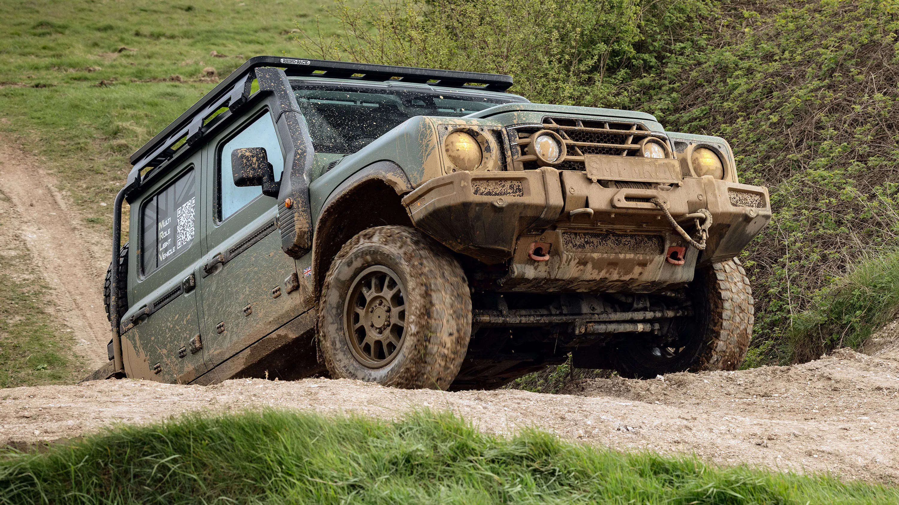 Green off-road vehicle with mud splatters climbing a dirt hill with grass and bushes in the background.