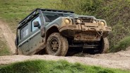 Green off-road vehicle with mud splatters climbing a dirt hill with grass and bushes in the background.