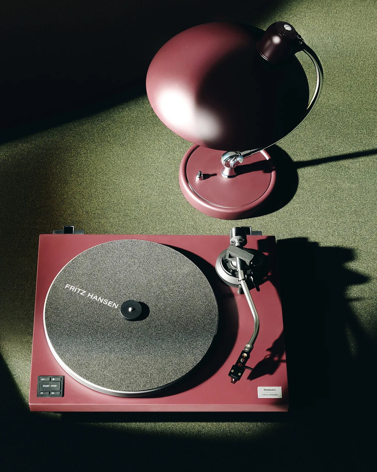 Burgundy Technics turntable with gray felt mat labeled "Fritz Hansen" next to matching burgundy desk lamp on green carpet.