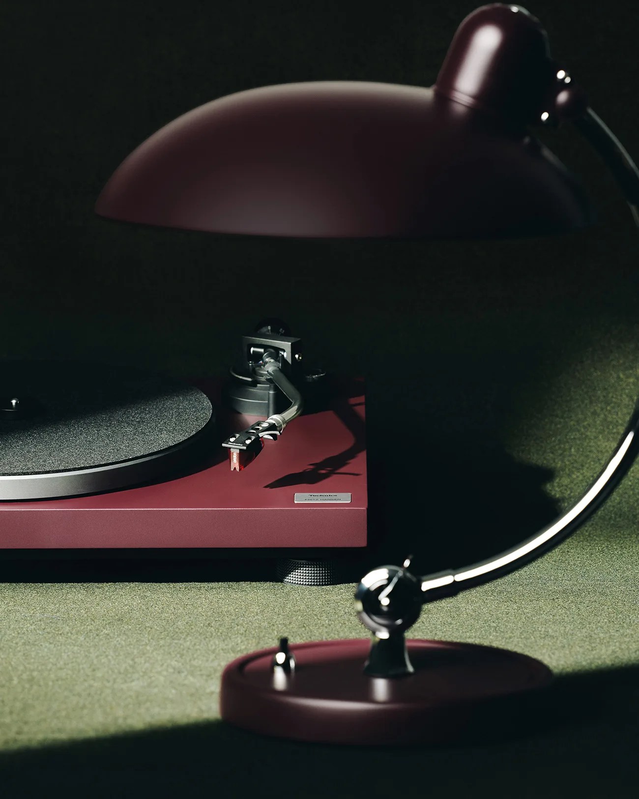 Burgundy Technics turntable with tonearm and a matching burgundy desk lamp on a green surface.