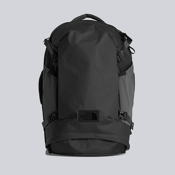Black The North Face backpack with top handle and side compression straps on a gray background.
