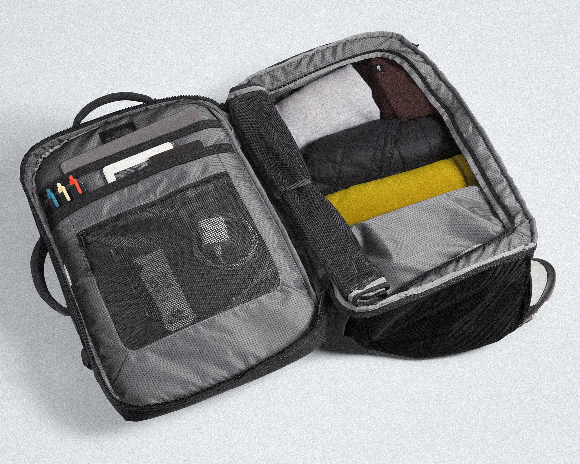 Open black travel backpack with organized compartments holding folded clothes, pens, and electronic accessories.