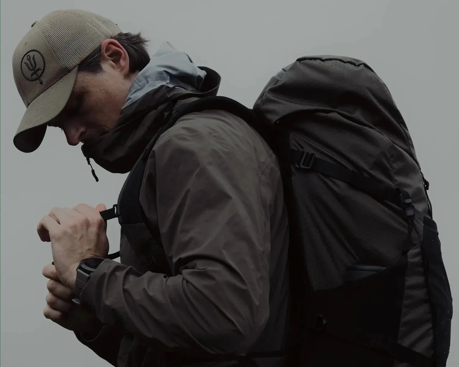 Man wearing a beige cap and dark jacket adjusting straps on a large black backpack.