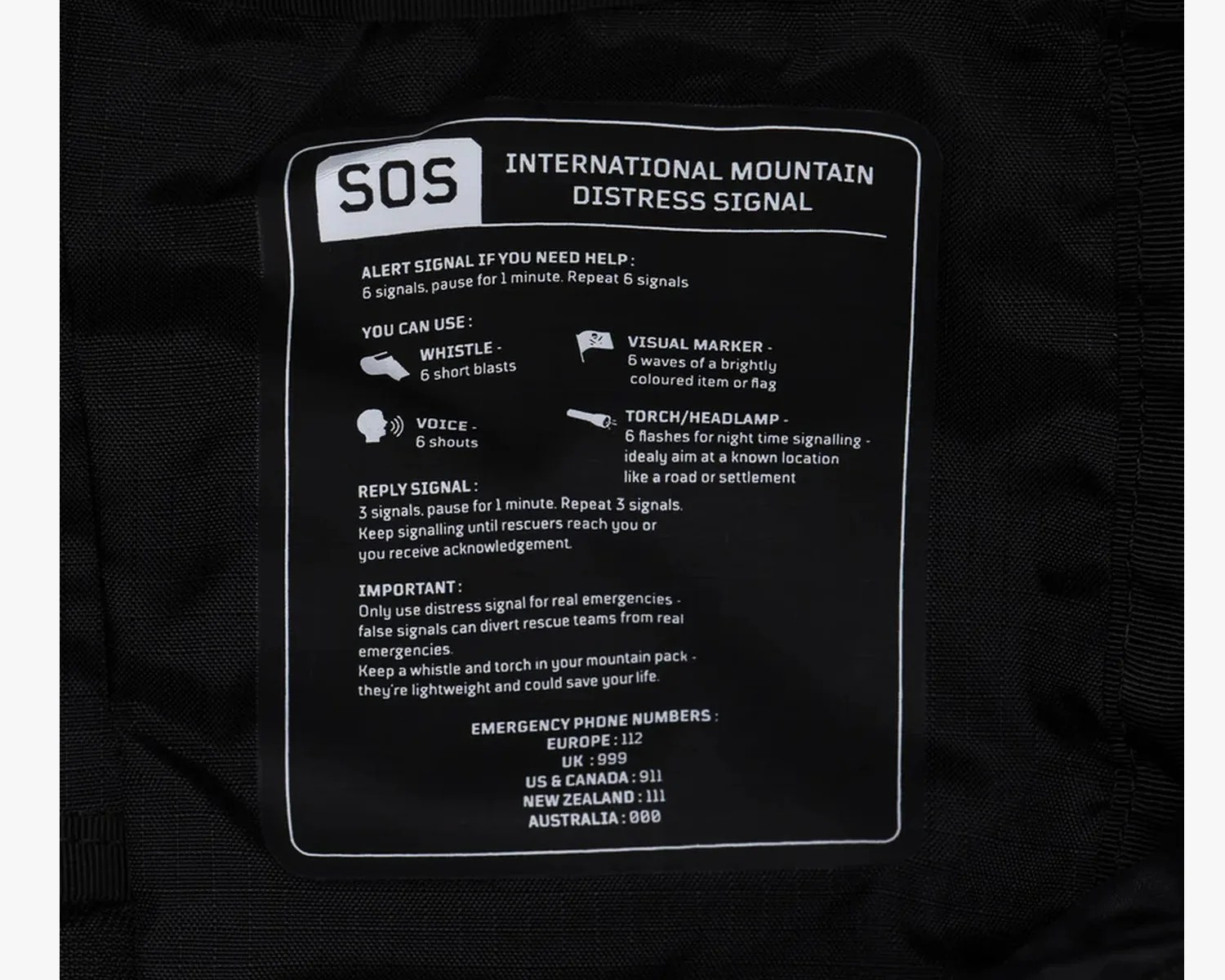 Black fabric label with white text explaining the international mountain distress signal SOS and emergency phone numbers.