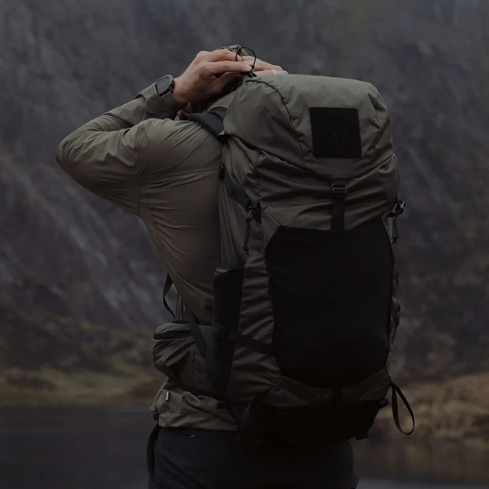 Olive green and black hiking backpack worn by a person adjusting the top strap outdoors.