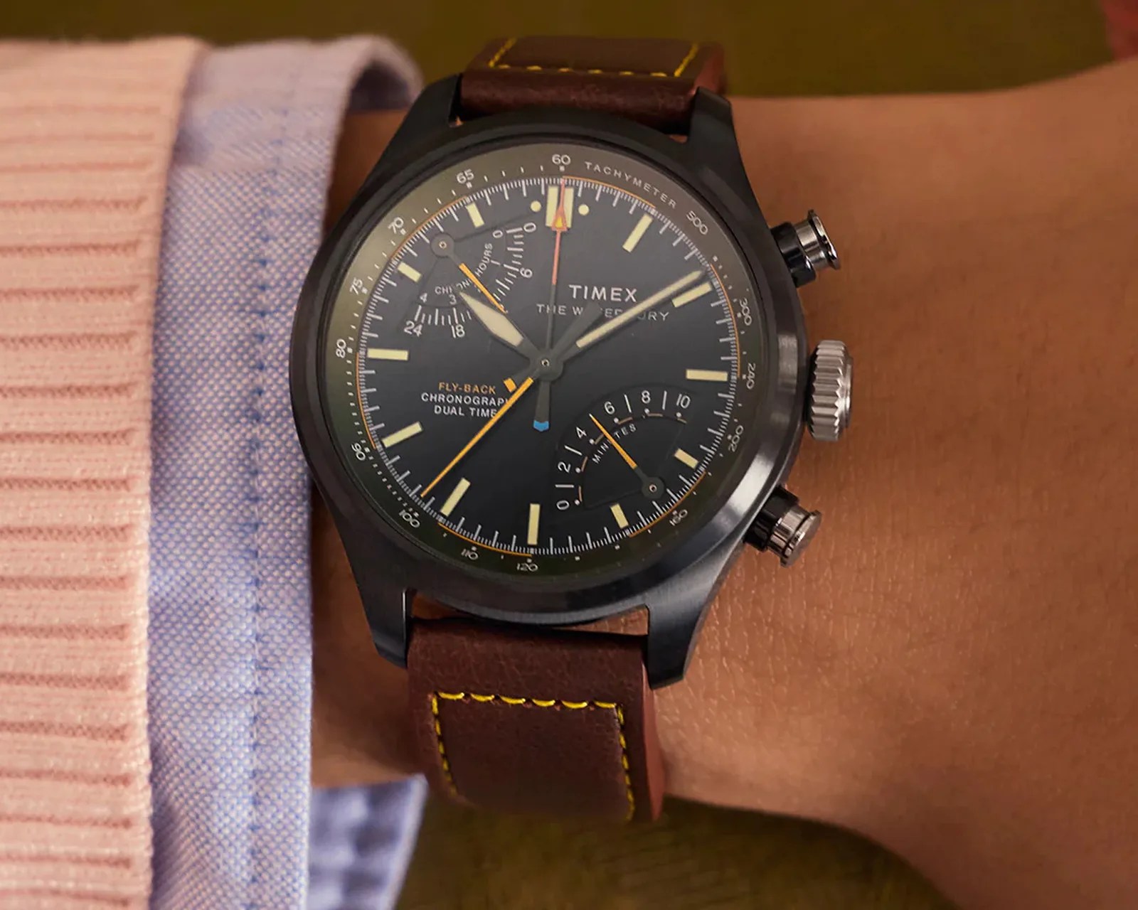 Black Timex chronograph watch with tachymeter and brown leather strap with yellow stitching on a wrist.