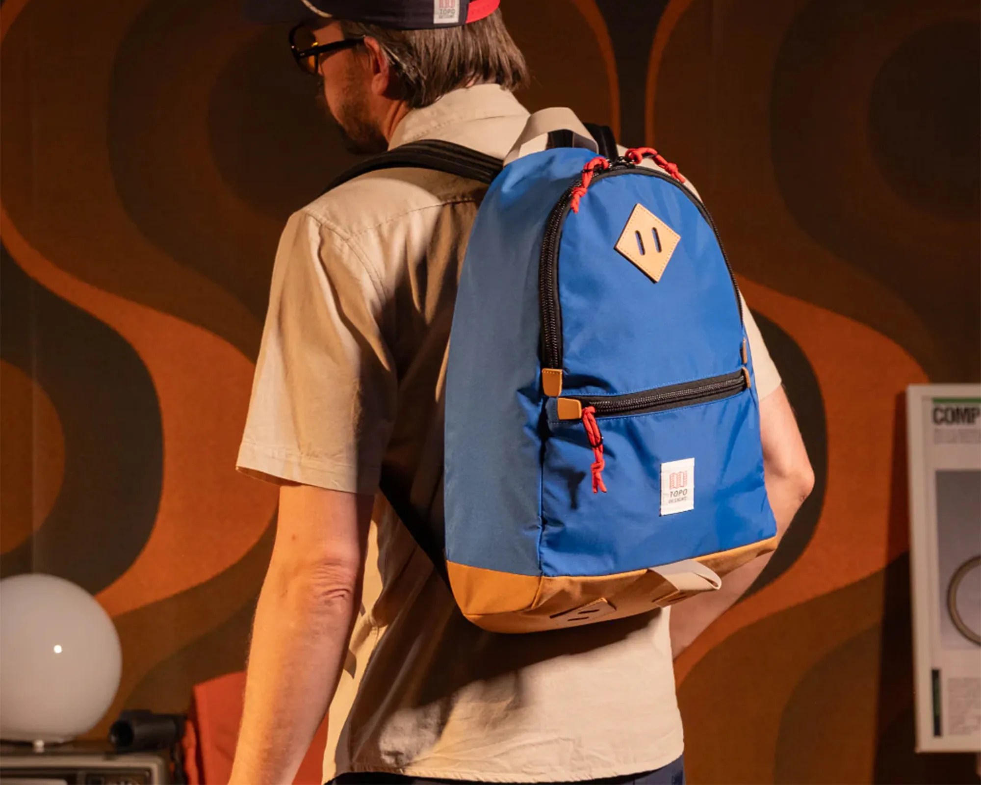 Blue and tan backpack with red zipper pulls worn by a person in a beige shirt.