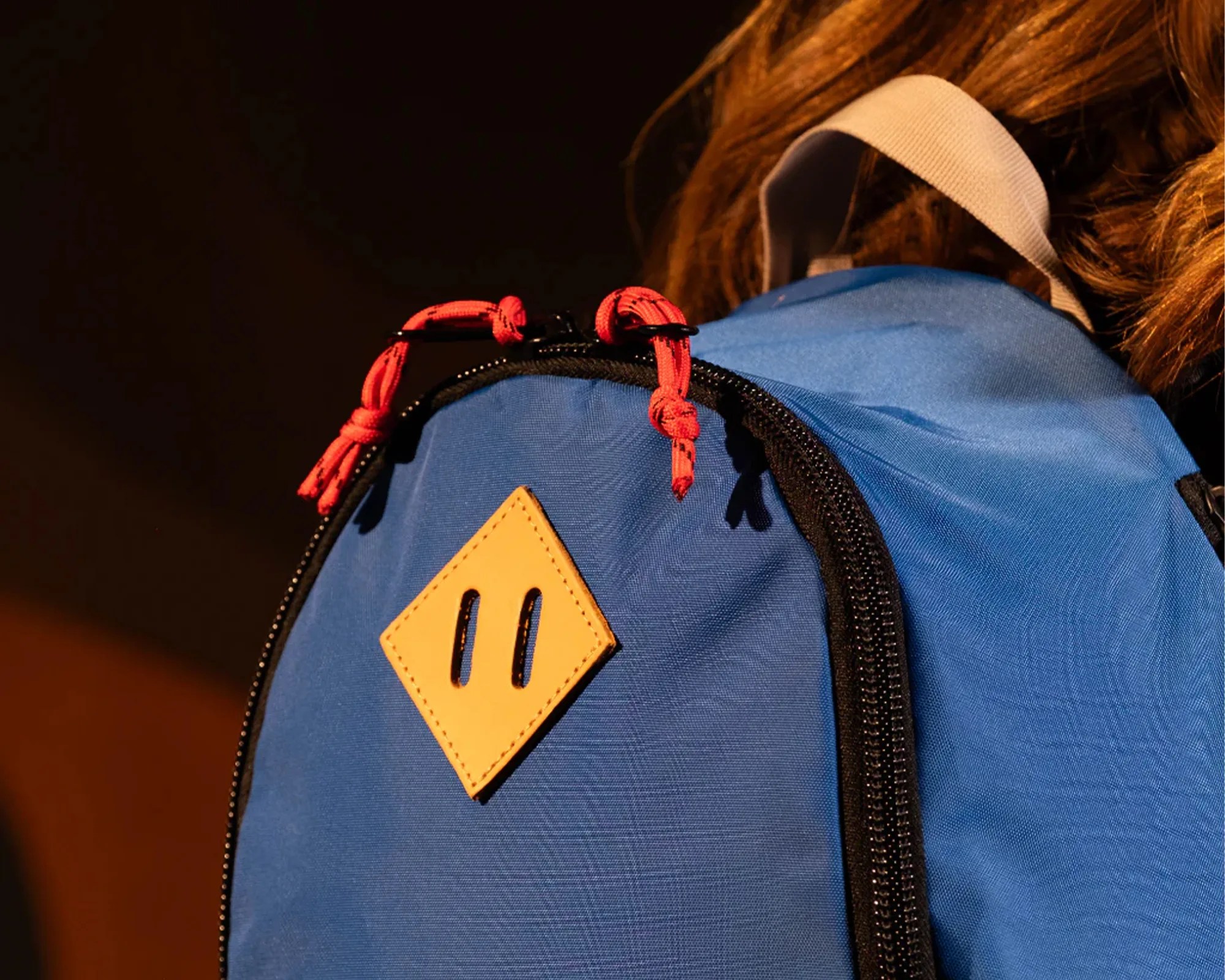 Blue backpack with black zipper, red zipper pulls, and a tan diamond-shaped leather patch.