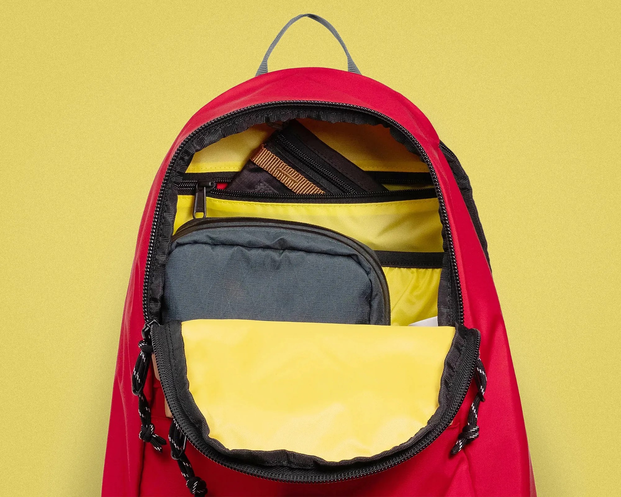 Red backpack with yellow interior and black zipper, partially open showing a black and gray pouch inside.