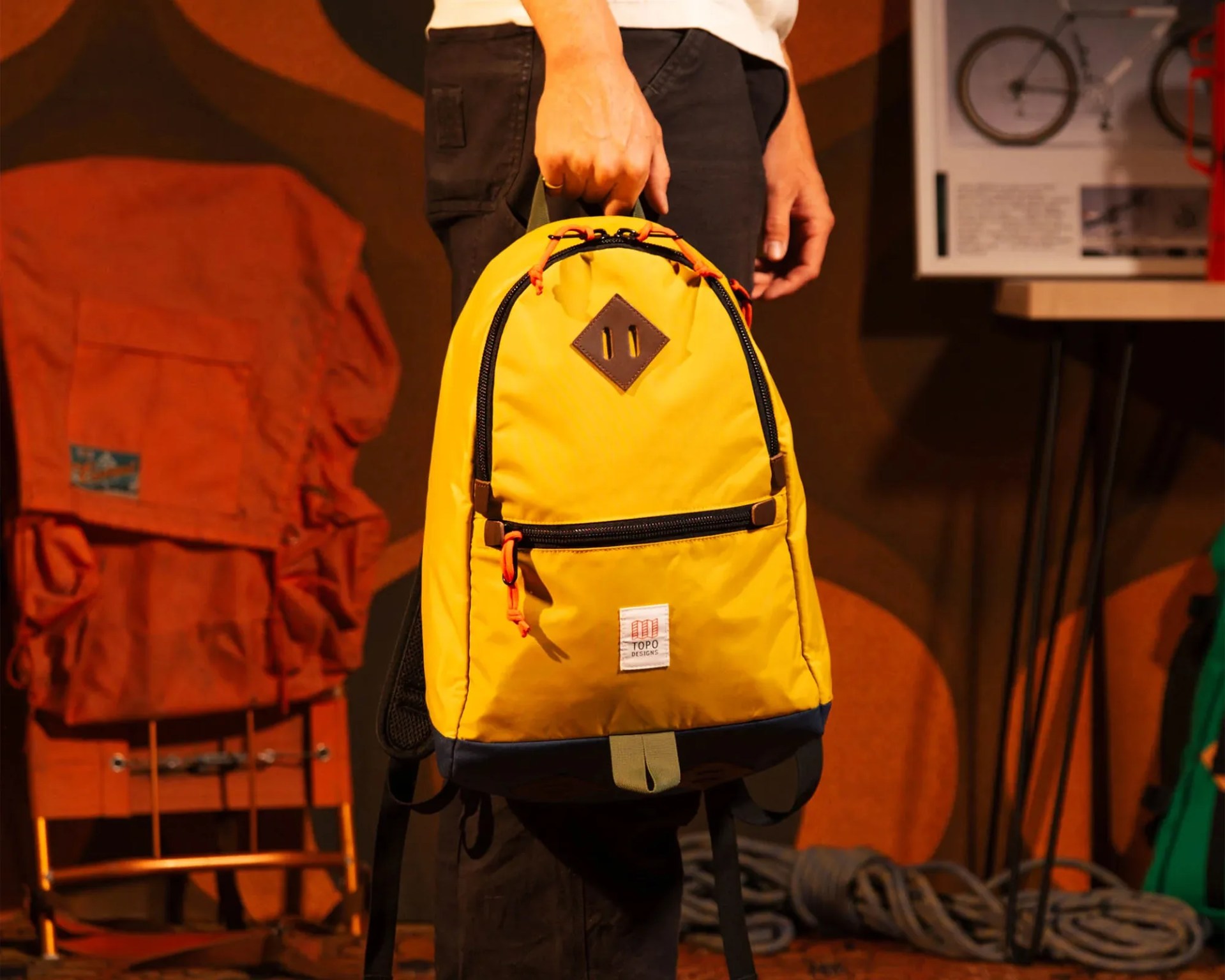 Person holding a yellow and navy Topo Designs backpack with black zippers and orange zipper pulls.