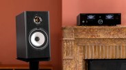 Black bookshelf speaker with white woofer cone on a stand next to a marble fireplace with a black audio receiver on the mantel.