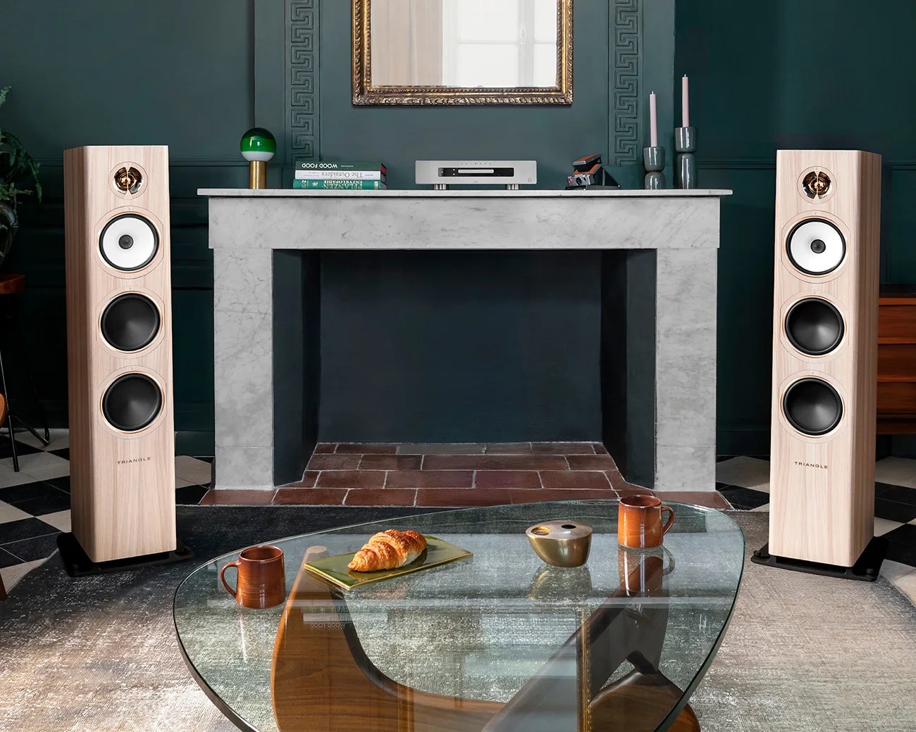 Pair of light wood floor-standing Triangle speakers with white and black drivers flanking a gray marble fireplace in a living room.