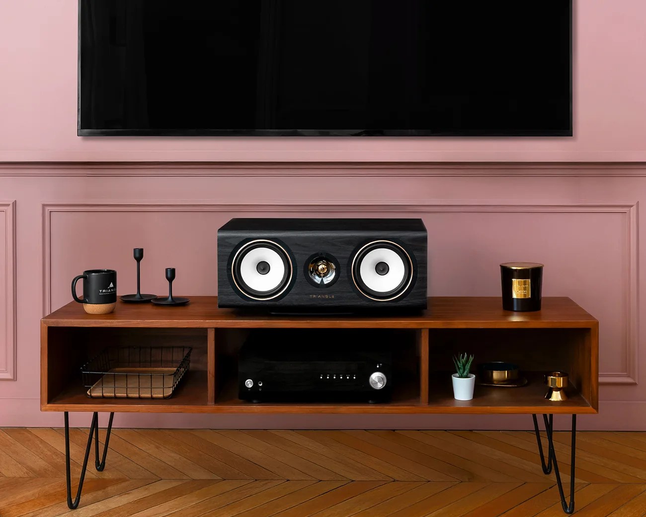 Black center speaker with white cones on a wooden media console with black metal legs against a pink paneled wall.