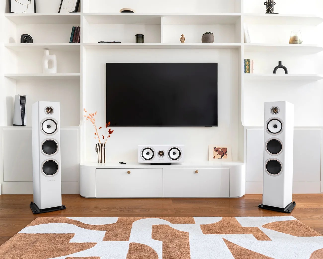 White floor-standing speakers and a matching center speaker below a wall-mounted flat-screen TV in a modern white entertainment unit.