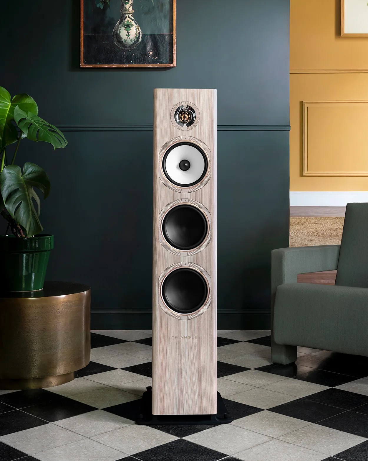 Tall light wood floor-standing speaker with three drivers on a black and white checkered floor.