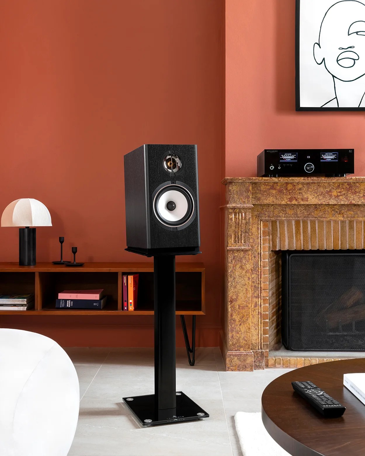 Black bookshelf speaker with white cone woofer on a black metal stand in a living room.