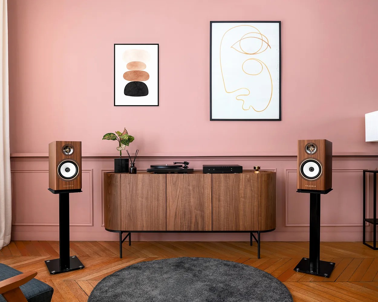 Pair of wooden bookshelf speakers on black stands flanking a wooden sideboard with audio equipment against a pink wall.