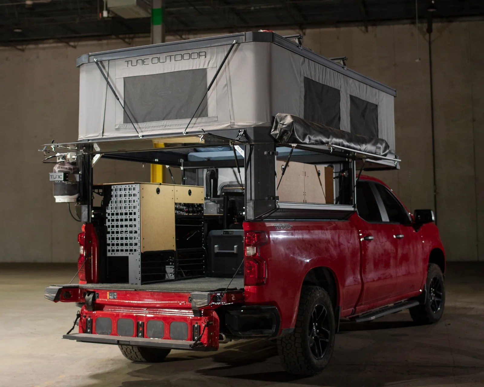Red pickup truck with a gray pop-up camper tent and storage units in the truck bed inside a warehouse.