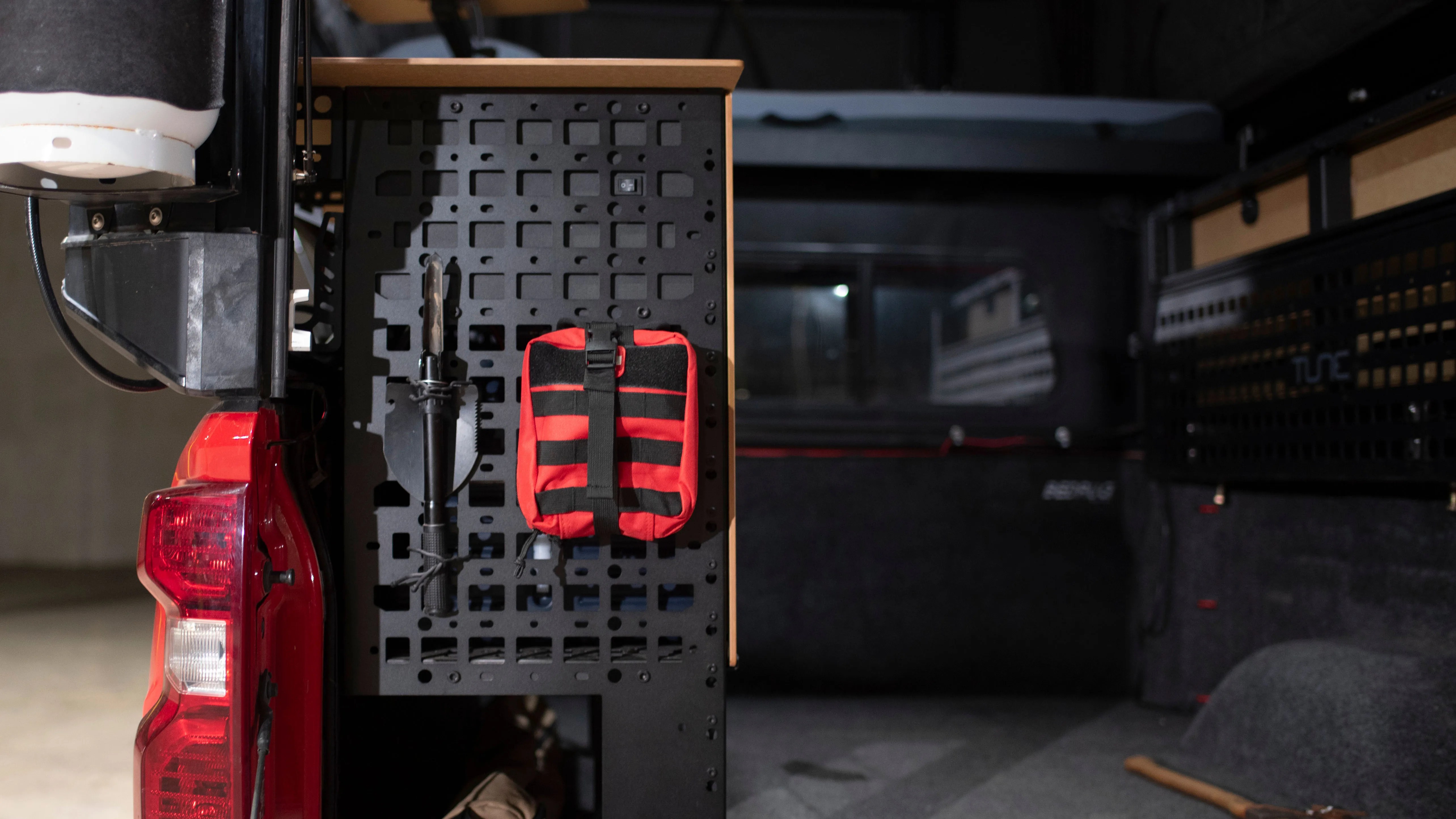 Black metal pegboard mounted inside a vehicle with a black-handled shovel and a red first aid kit attached.