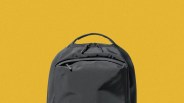 Black backpack with top handle and front zippered pocket against a yellow background.