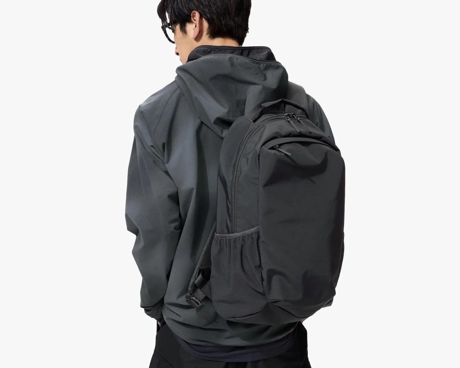 Black backpack with multiple zippered compartments worn by a person in a dark gray hooded jacket.