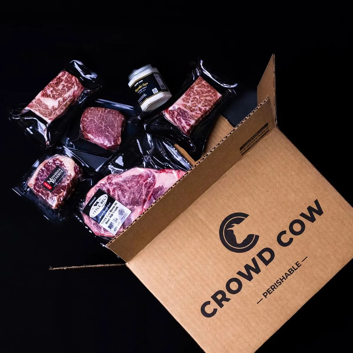 Open cardboard box labeled "Crowd Cow Perishable" containing various vacuum-sealed cuts of marbled beef and a jar of seasoning.