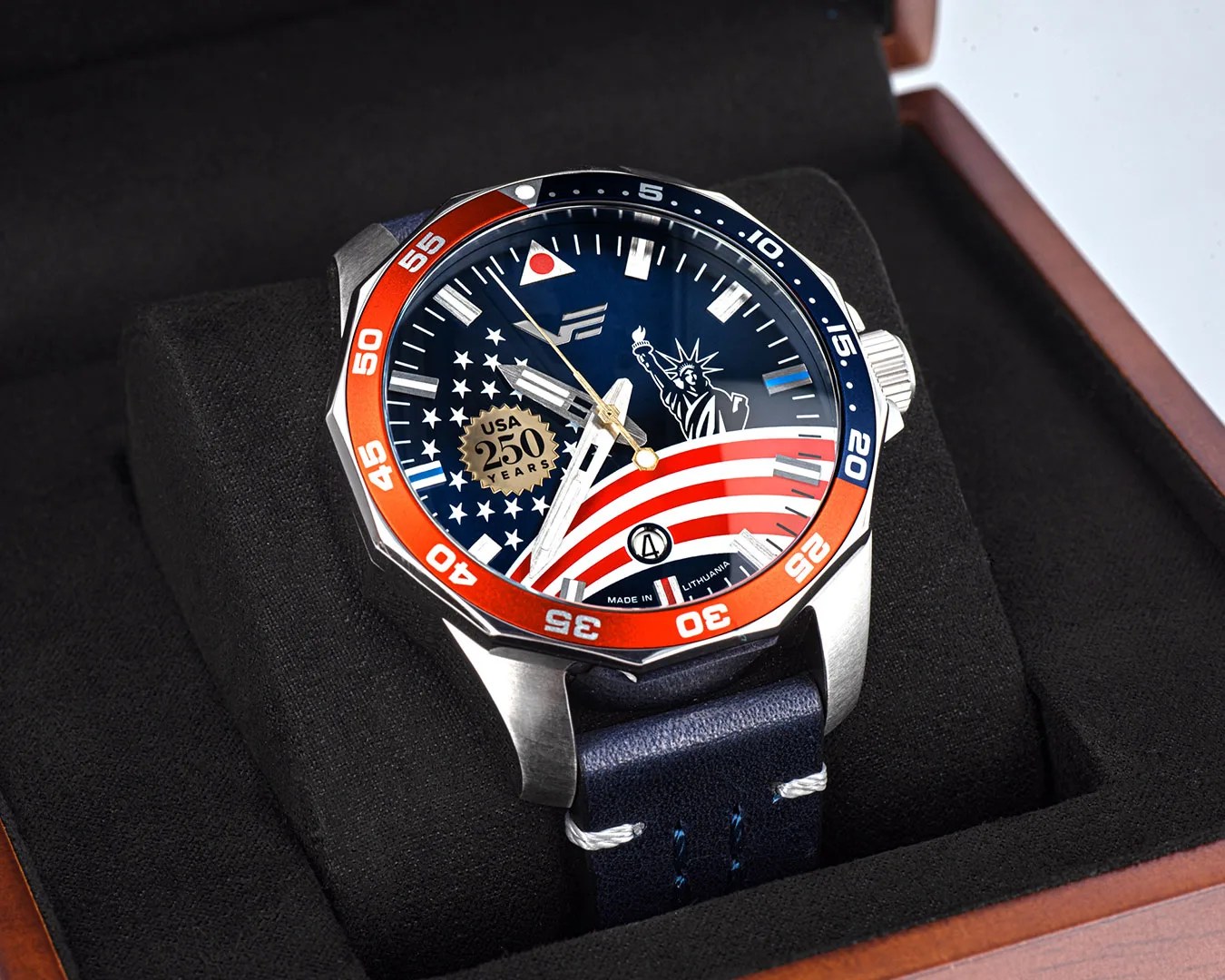 Silver watch with red and blue bezel, American flag and Statue of Liberty design on dial, and blue leather strap in a black cushion.
