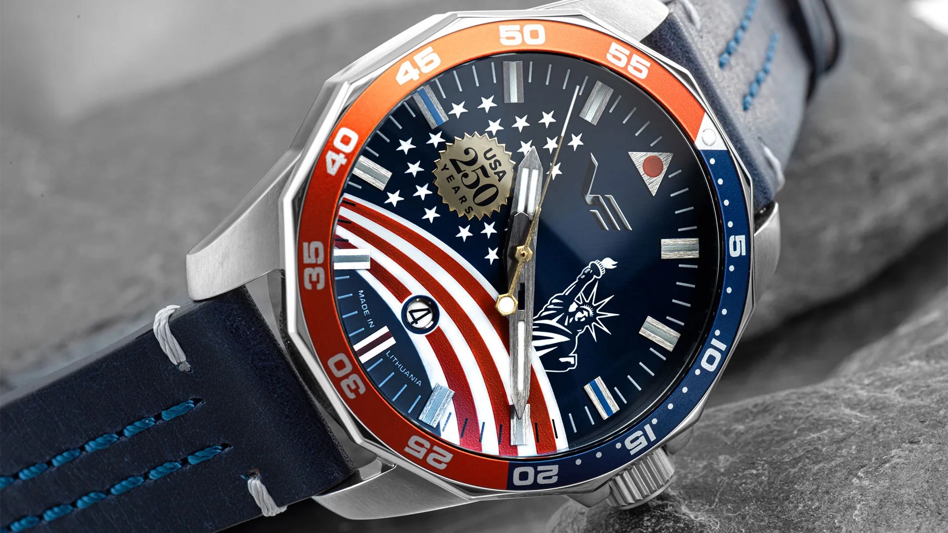Close-up of a wristwatch with a blue dial featuring the Statue of Liberty, American flag stripes, stars, and a "USA 250 Years" emblem, with an orange and blue bezel and a blue leather strap.