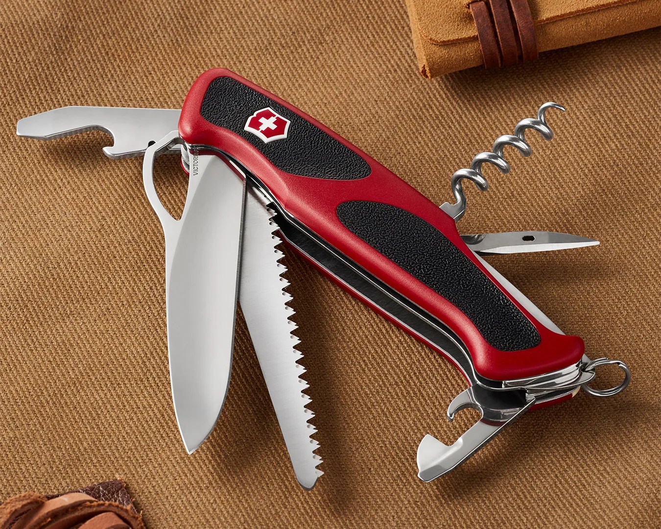 Red and black Swiss Army knife with multiple tools extended, including a blade, saw, corkscrew, and bottle opener, on a brown fabric surface.