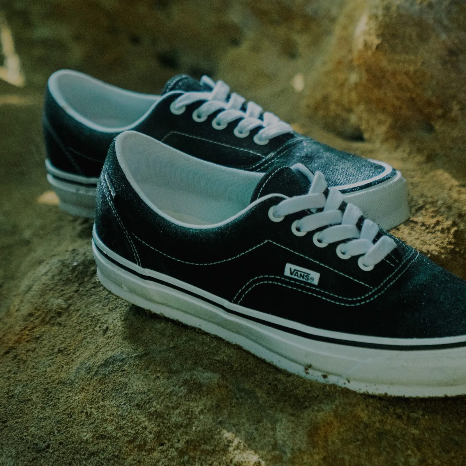 Pair of black Vans sneakers with white laces and soles on a rocky surface.