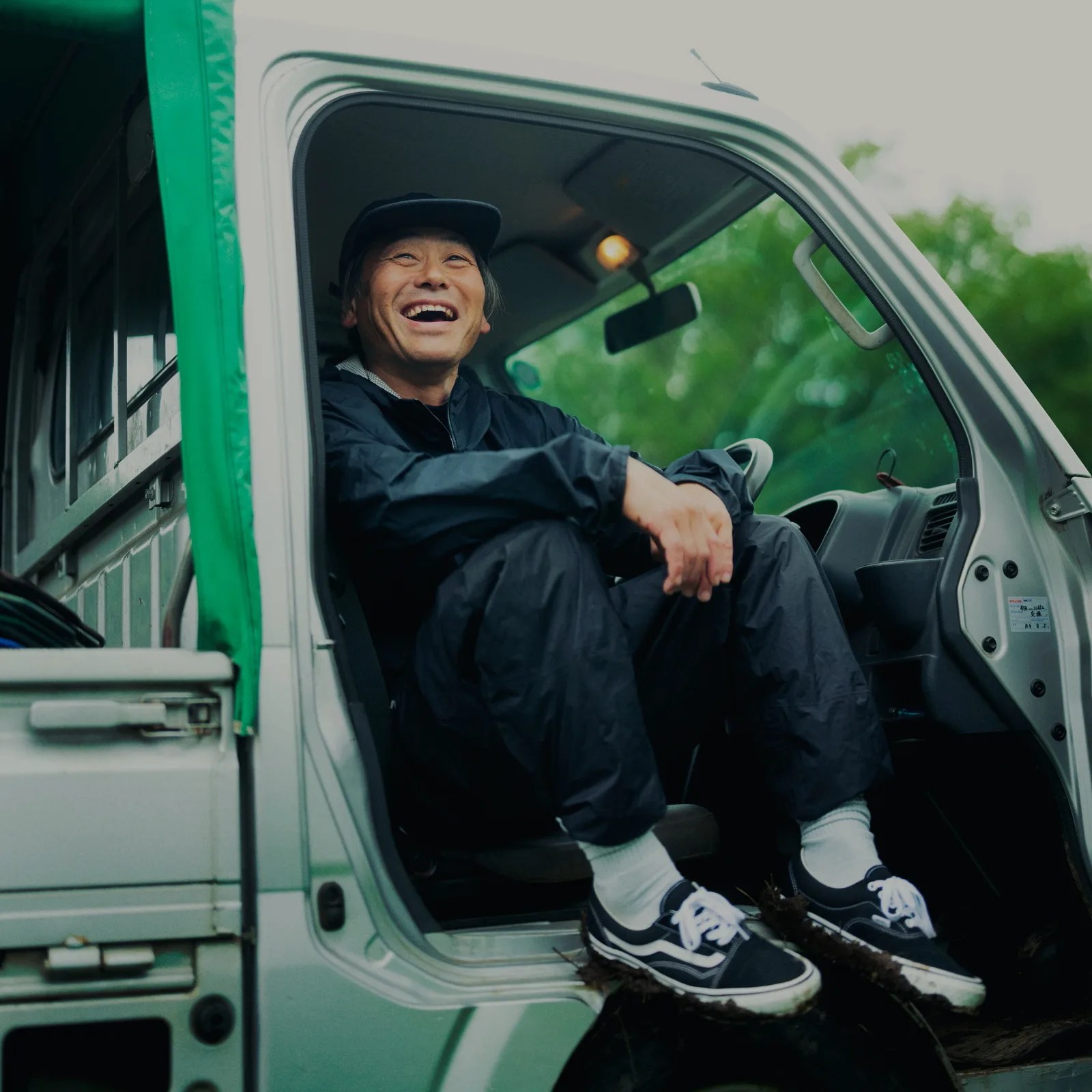 Man in black rain suit and cap sitting in the driver's seat of a white truck with green tarp.
