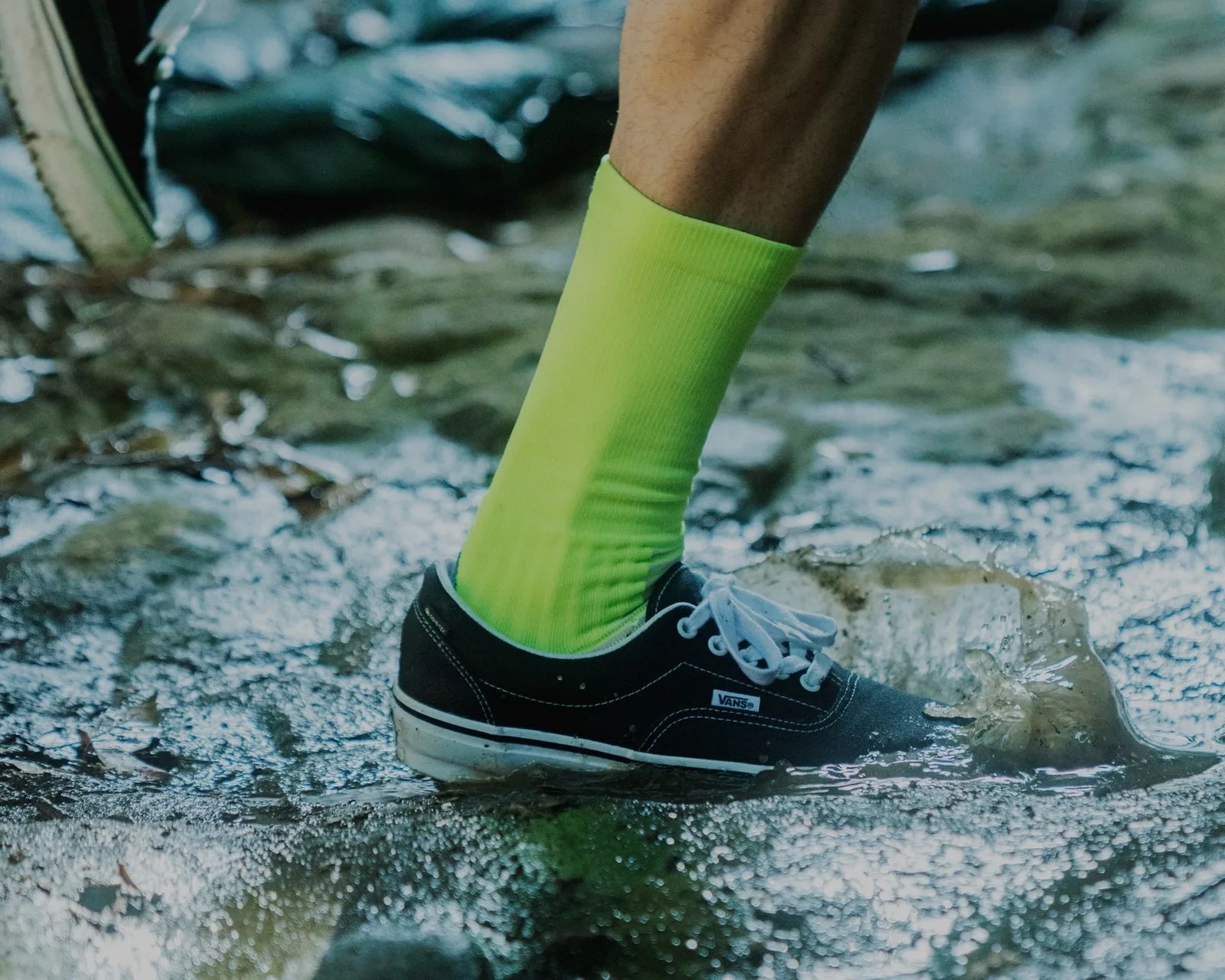 Black Vans sneaker with white laces and neon green sock stepping in a water puddle.
