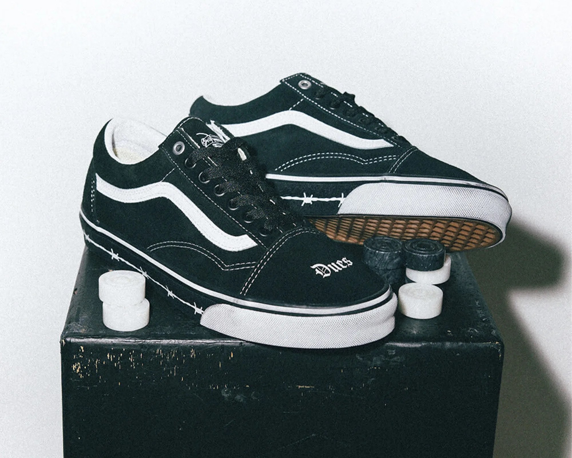 Black Vans sneakers with white side stripe and "Dues" text on toe, placed on a black box with skateboard wheels.