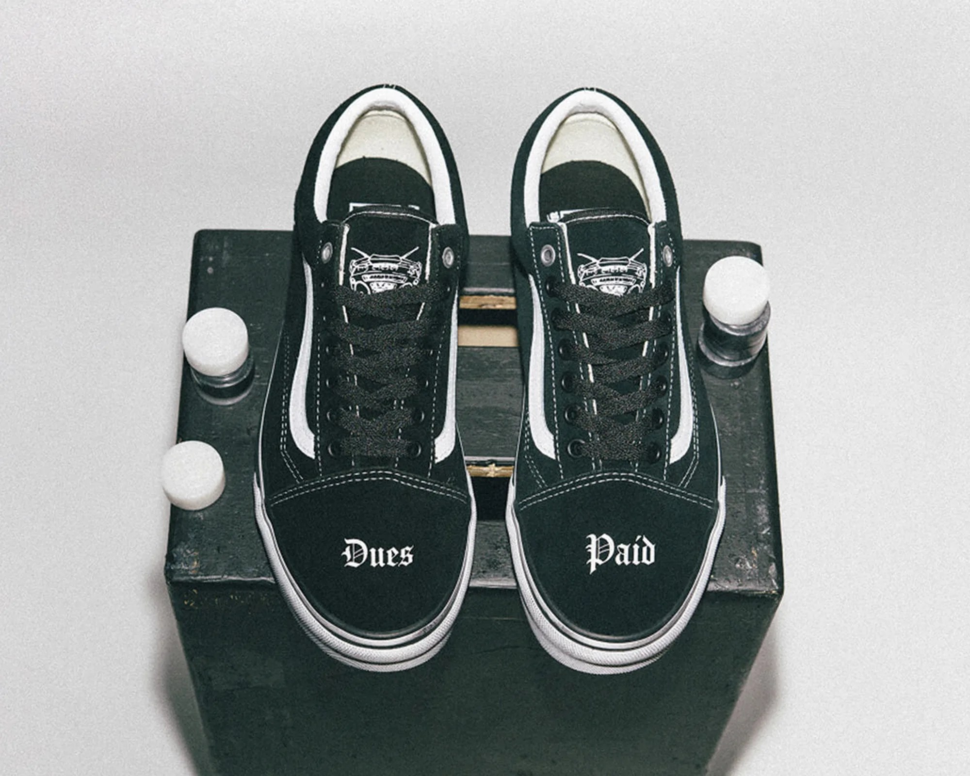 Pair of black Vans sneakers with white side stripes and "Dues Paid" text on the toes, placed on a black box.