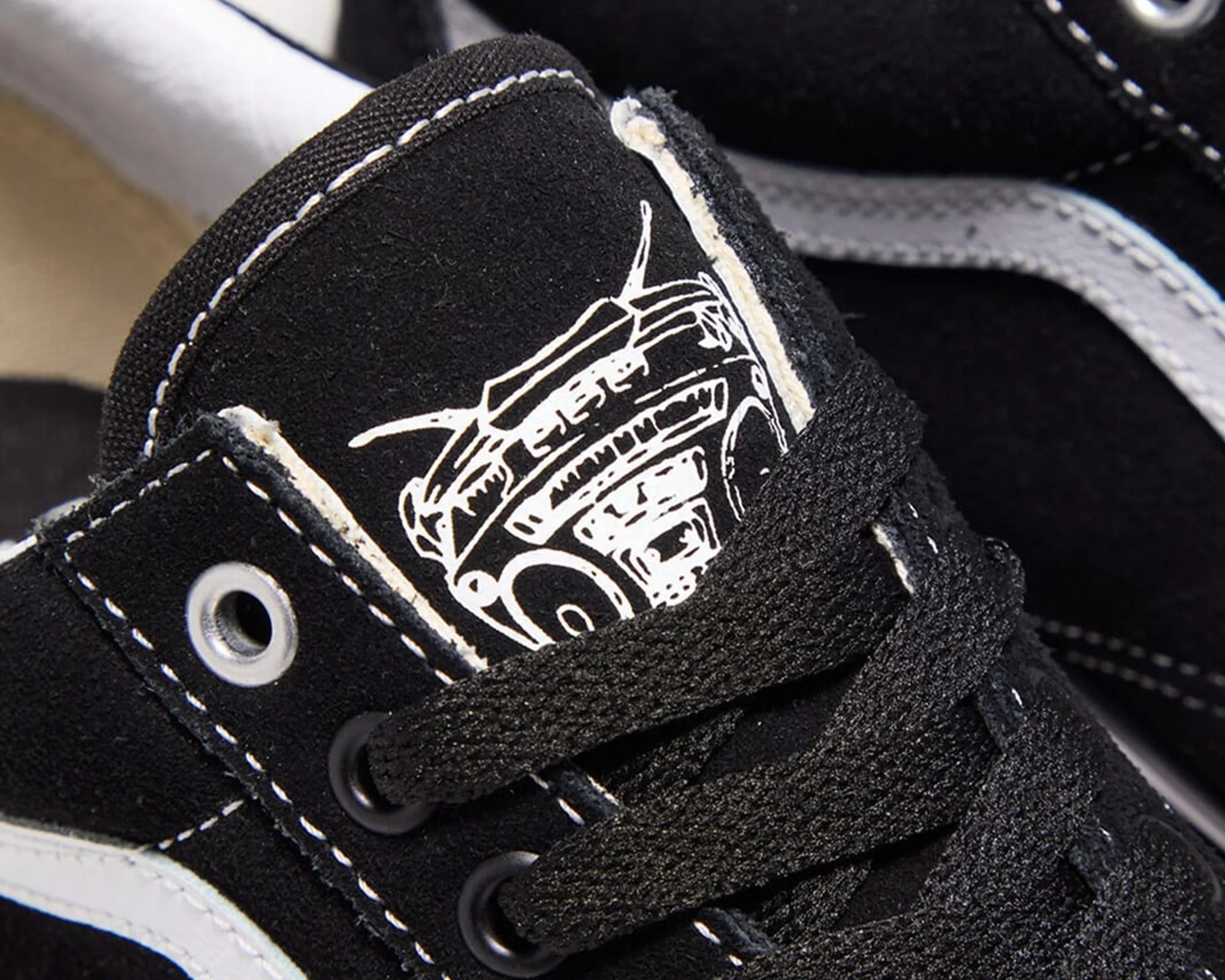 Close-up of black suede sneaker tongue with white graphic and black laces threaded through metal eyelets.