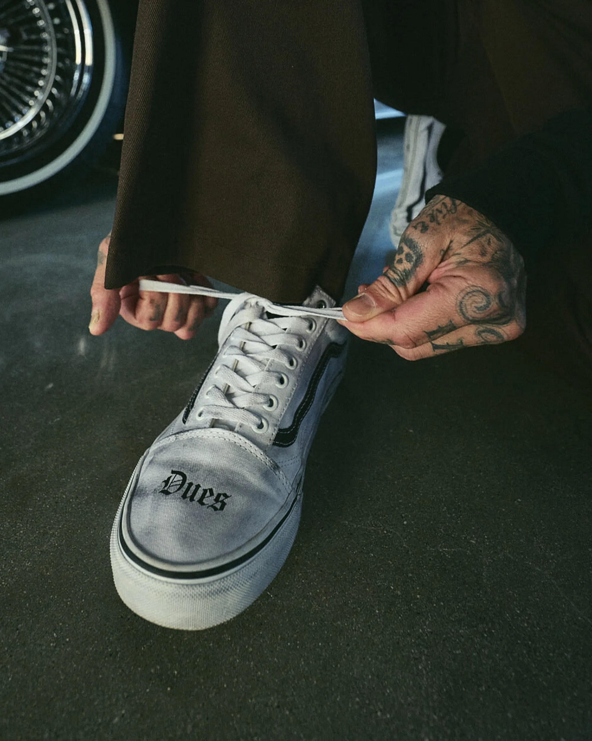 White canvas sneaker with black stripe and "Dues" text being tied by tattooed hands.