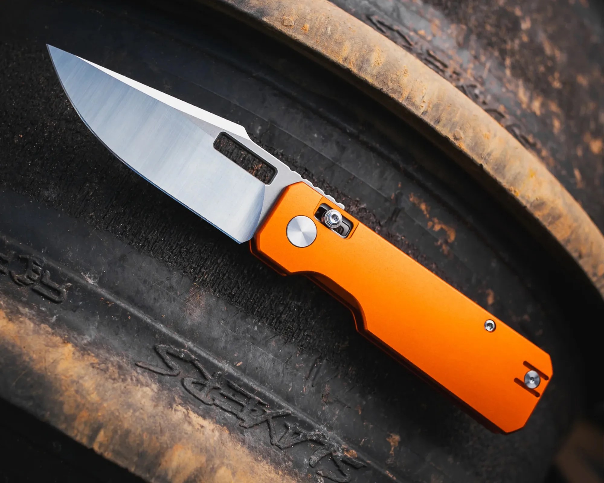 Knife with a silver blade and orange handle resting on a worn black tire.
