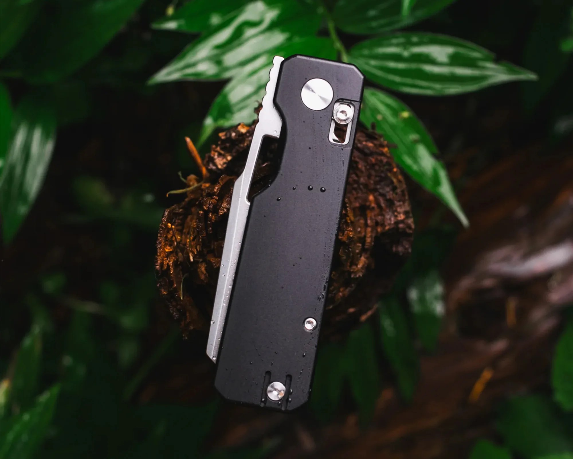 Black and silver folding knife with screws and a thumb stud, resting on a tree stump with green leaves in the background.