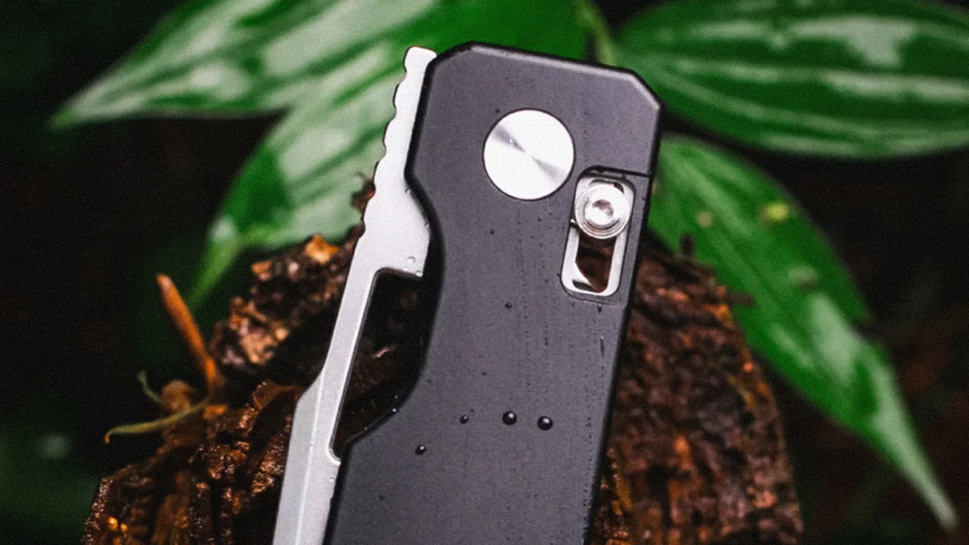 Black and silver multitool with a circular pivot and a small screw, resting on soil with green leaves in the background.